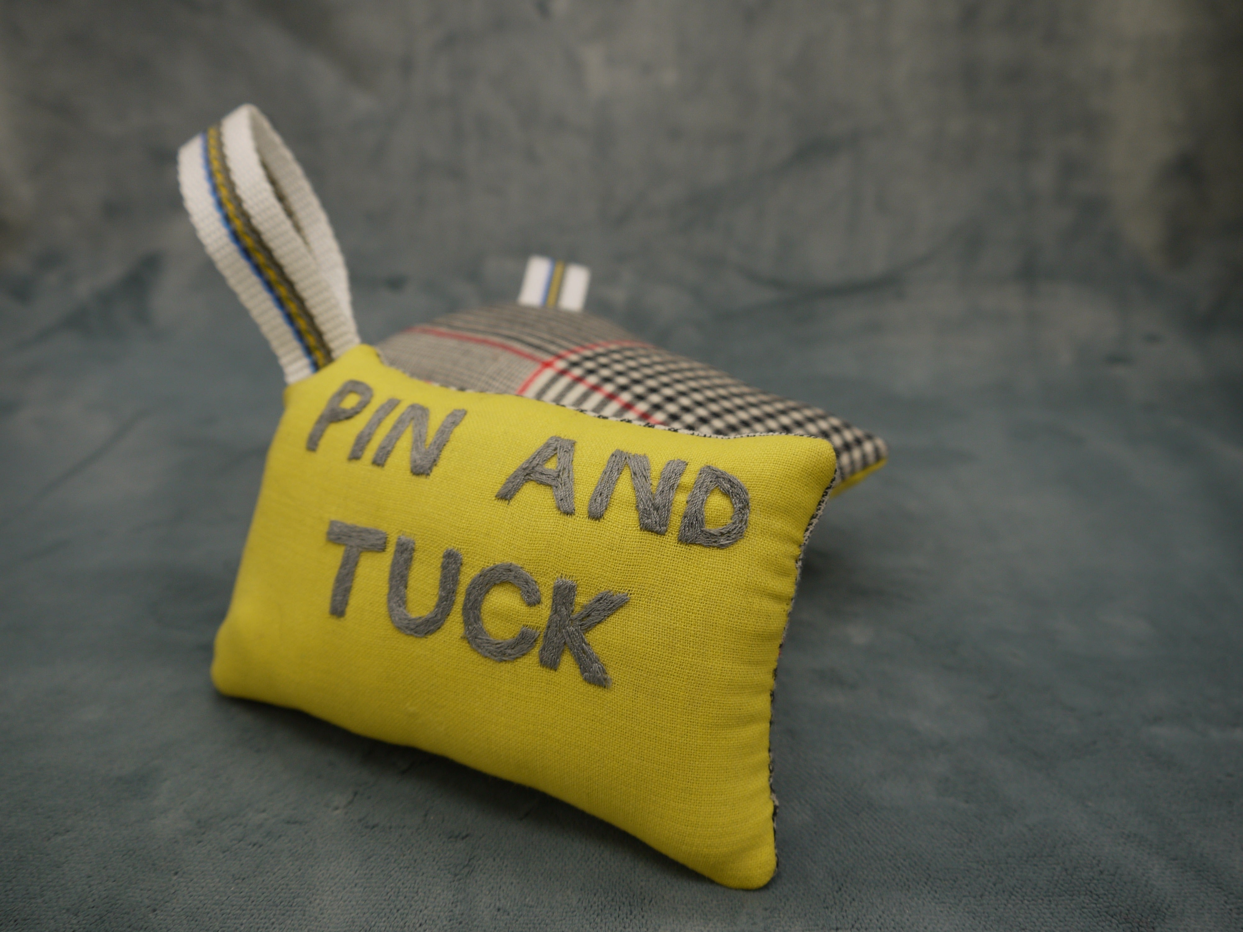 Pin cushion in yellow and grey wool, superfine 120, Pin and Tuck Embroidered by hand in grey on a yellow background, Nylon wriststrap with yellow/blue go stripe, Prince of Wales wool backing in white and black