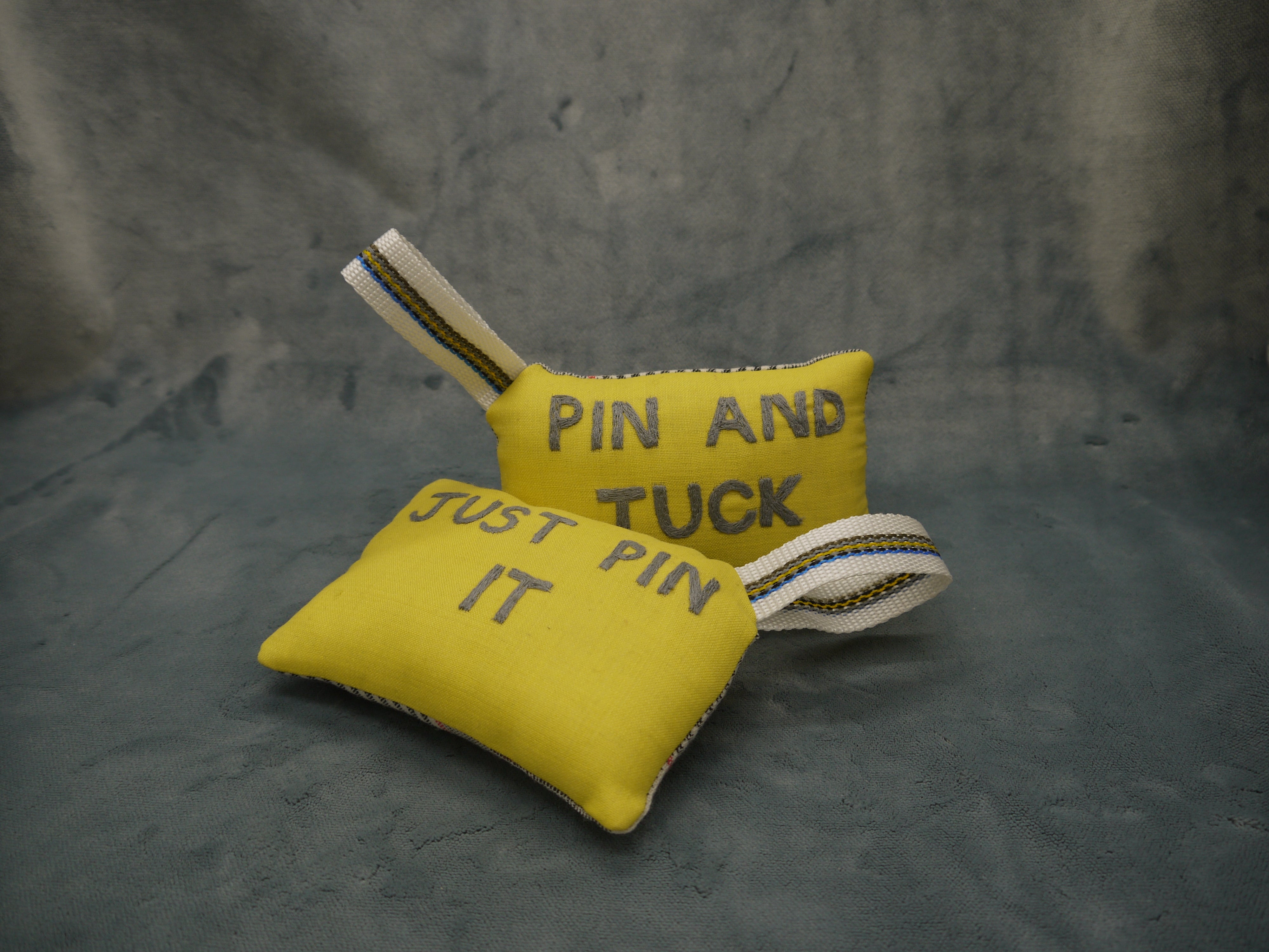 Pin cushion in yellow and grey wool, superfine 120, Just Pin It Embroidered by hand in grey on a yellow background, Nylon wriststrap with yellow/blue go stripe, Prince of Wales wool backing in white and black.PIn and Tuck  pincushion visible in the background