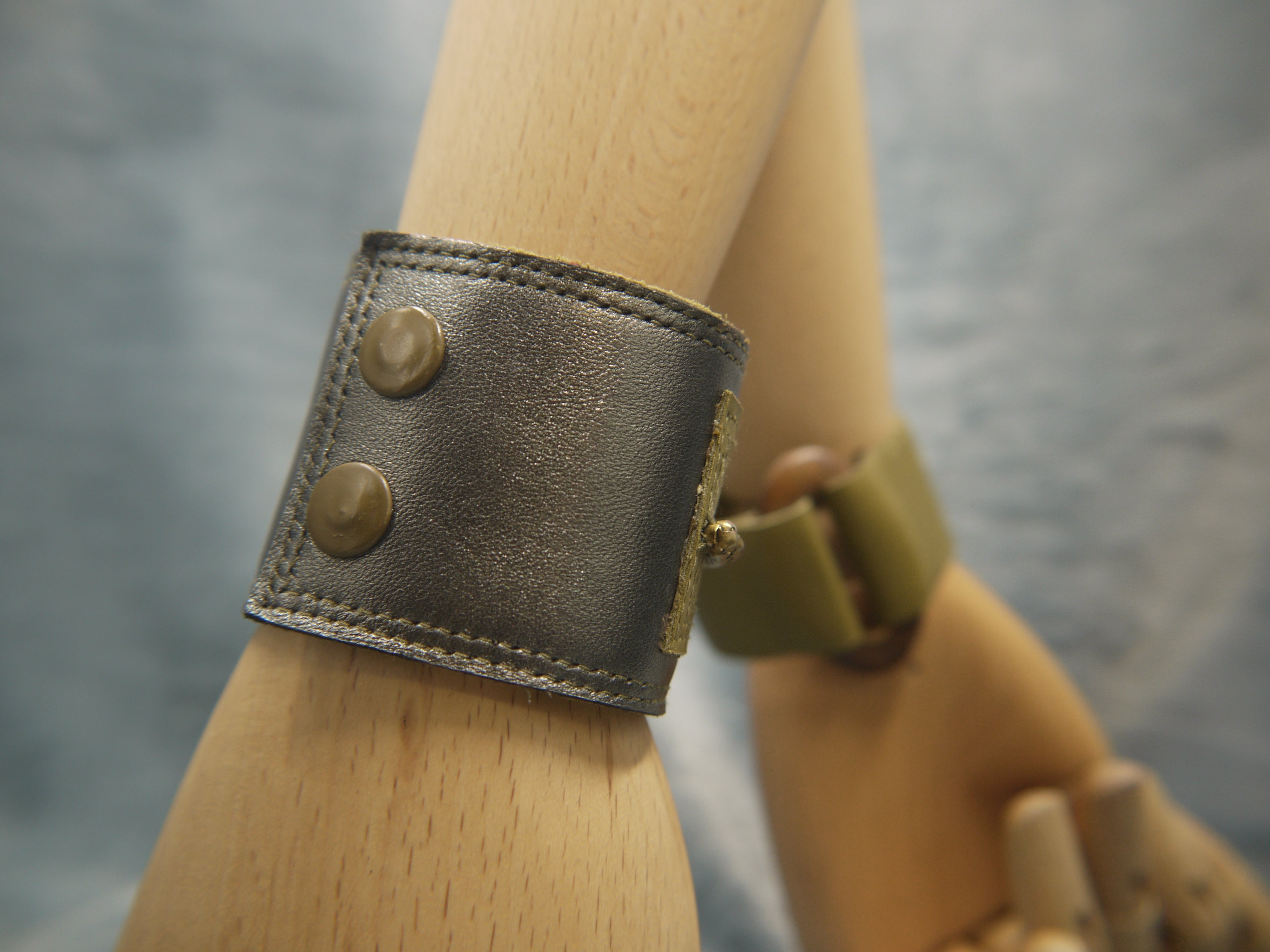 Leather cuff in Gunmetal metallic leather and moss green leather central rectangle which has repeat stich detail which frames the rectangle. A central brass chain is the focal point of the cuff, sewn central across the moss green rectangle. Moss Green/Oval wood cuff seen in the background